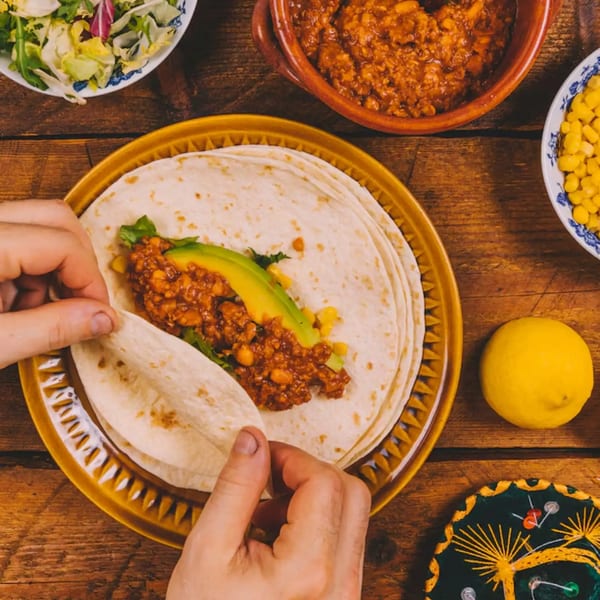 Hands preparing a Mexican taco with meat, avocado, and corn on a wooden table.