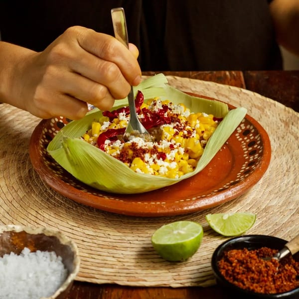A hand forks into a Mexican esquites dish with corn, cheese, chili, and lime.